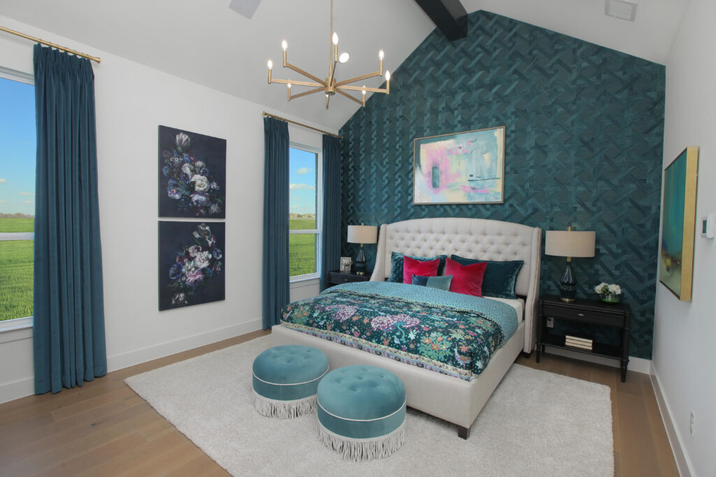 Modern bedroom in the Painted Tree community, with a patterned teal accent wall, upholstered bed, floral bedding, teal ottomans, gold chandelier, and large windows with blue curtains—your perfect new home for sale in McKinney TX.