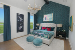 Modern bedroom in the Painted Tree community, with a patterned teal accent wall, upholstered bed, floral bedding, teal ottomans, gold chandelier, and large windows with blue curtains—your perfect new home for sale in McKinney TX.