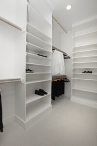 A modern walk-in closet with white shelves, hanging rods, and neatly arranged black shoes and white shirts—perfect for a Drees Custom Homes residence in the Painted Tree community of McKinney TX.