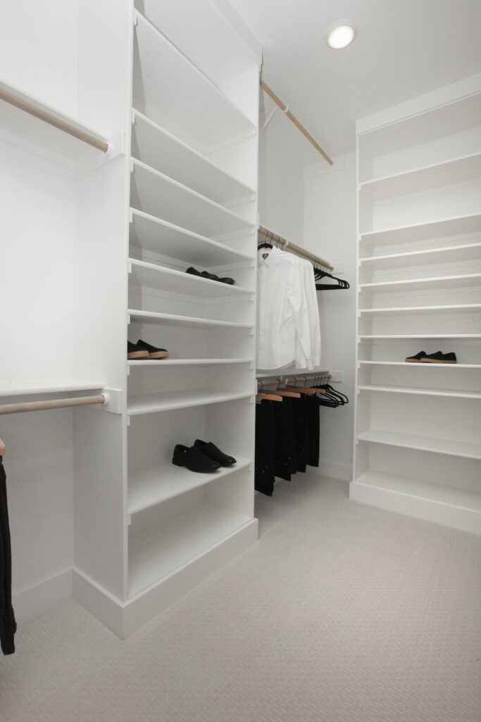 A modern walk-in closet with white shelves, hanging rods, and neatly arranged black shoes and white shirts—perfect for a Drees Custom Homes residence in the Painted Tree community of McKinney TX.