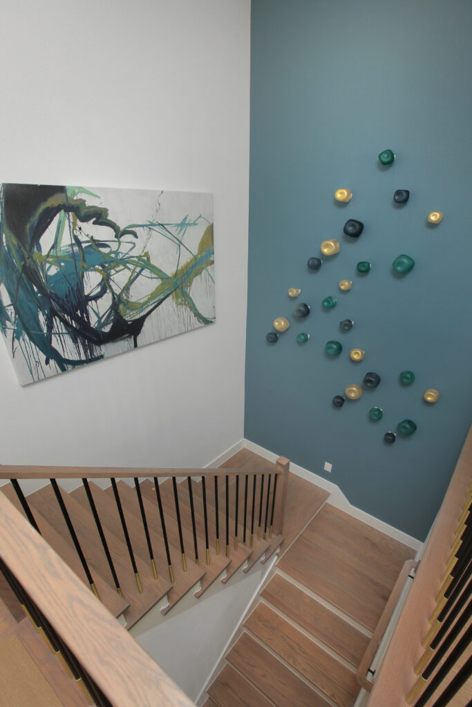 A modern staircase with wooden steps and railings, abstract artwork on one wall, and decorative wall-mounted objects on a teal accent wall in a beautiful new home for sale in the Painted Tree community, McKinney TX.