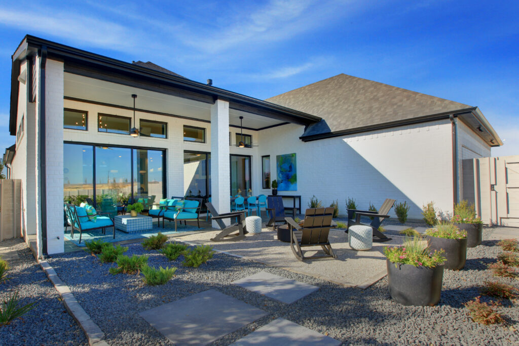 Modern backyard patio with outdoor seating, potted plants, and gravel landscaping highlights this new home for sale in McKinney TX by Drees Custom Homes, featuring large sliding glass doors leading into a white brick house under a blue sky.