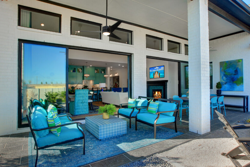 Modern outdoor patio with blue cushioned seating and table, overlooking an open living room with contemporary decor through large sliding glass doors in the Painted Tree community by Drees Custom Homes.