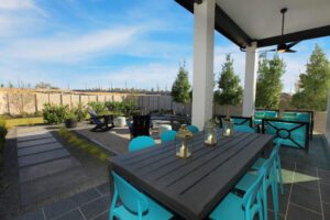 Modern backyard patio at a new home for sale by Drees Custom Homes in McKinney TX, featuring a long dining table, turquoise chairs, lanterns, outdoor seating area, and landscaped garden under a partly cloudy sky.
