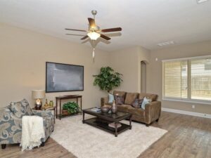 A living room in Painted Tree McKinney features a brown sofa, patterned armchair, glass coffee table, area rug, wall art, potted plant, and ceiling fan. A large window fills the space with natural light—perfect for homes for sale in McKinney Texas.