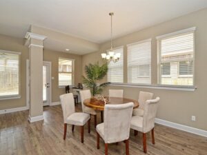 A dining room with a round wooden table, six upholstered chairs, hardwood floors, beige walls, large windows, and a chandelier offers elegant comfort—perfect for gatherings in Painted Tree McKinney homes. A potted plant adds a touch of nature in the corner.