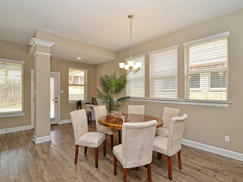 A dining room with a round wooden table, six upholstered chairs, hardwood floors, beige walls, large windows, and a chandelier offers elegant comfort—perfect for gatherings in Painted Tree McKinney homes. A potted plant adds a touch of nature in the corner.