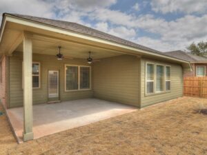 Covered backyard patio with concrete floor, ceiling fans, and green siding, overlooking a dry lawn and fenced area—perfect for enjoying evenings in Painted Tree McKinney.