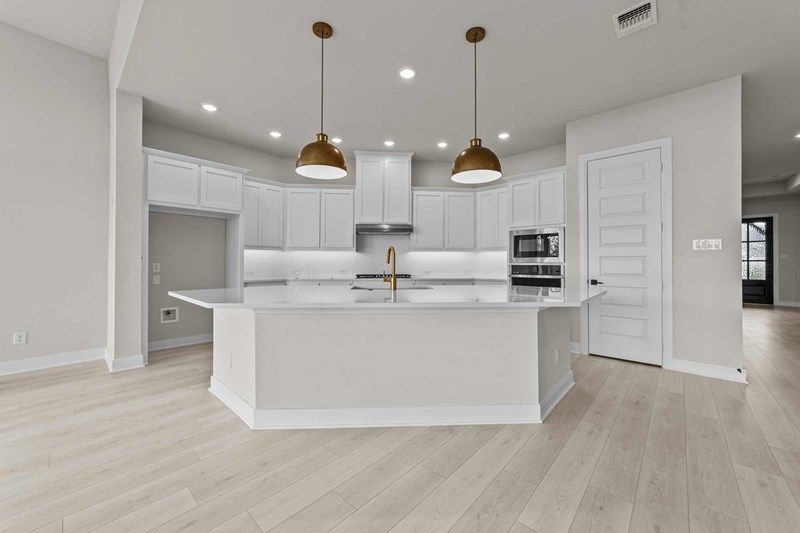 Modern kitchen with white cabinetry, stainless steel appliances, gold fixtures, two pendant lights, and light wood flooring—perfect for those seeking Painted Tree McKinney style or exploring homes for sale in McKinney, Texas.