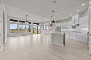 Open-concept kitchen and living area with white cabinets, light wood flooring, large windows, recessed lighting, and pendant lights over the island—perfect for those seeking Painted Tree homes for sale in McKinney.