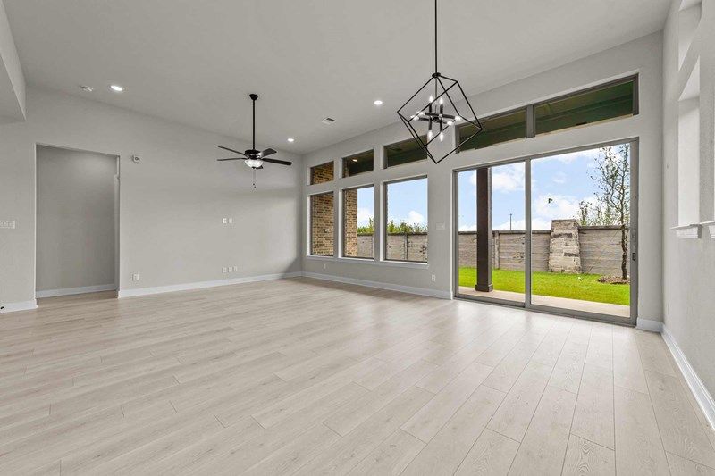Modern empty living room with light wood floors, large windows, sliding glass doors to a backyard, and white walls—perfect for those seeking Painted Tree McKinney homes for sale. The space features two sleek ceiling light fixtures.