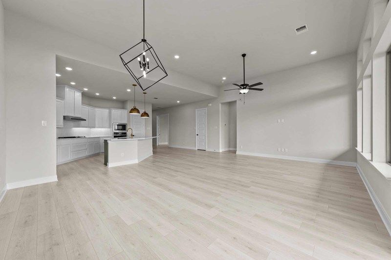 Open-plan room with light wood floors, white walls, modern kitchen with island, built-in oven, and pendant lights, plus large windows and a ceiling fan—perfect for those seeking homes for sale in McKinney Texas.