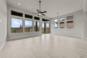 Spacious, empty living room with light wood flooring, large windows, ceiling fans, and glass door leading to a fenced backyard in Painted Tree McKinney—perfect for those searching for homes for sale in McKinney Texas.
