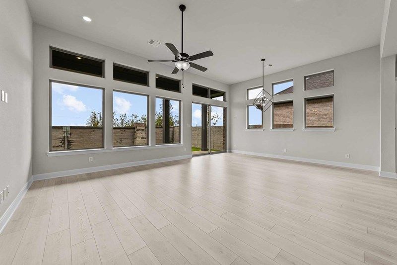 Spacious, empty living room with light wood flooring, large windows, ceiling fans, and glass door leading to a fenced backyard in Painted Tree McKinney—perfect for those searching for homes for sale in McKinney Texas.