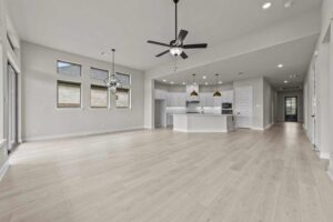 Spacious, empty open-concept living area in Painted Tree McKinney featuring light wood flooring, modern ceiling fans, large windows, and a white kitchen with island and pendant lights.
