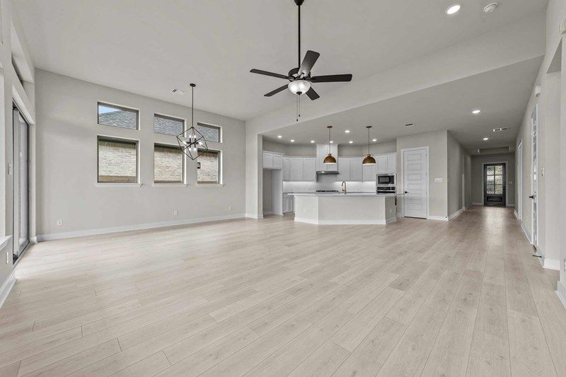 Spacious, empty open-concept living area in Painted Tree McKinney featuring light wood flooring, modern ceiling fans, large windows, and a white kitchen with island and pendant lights.