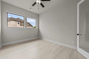 Bright empty room with light wood flooring, two windows showing a neighboring house, white walls, ceiling fan with light, and glass door—ideal for anyone seeking Painted Tree McKinney homes for sale.