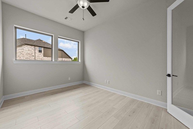 Bright empty room with light wood flooring, two windows showing a neighboring house, white walls, ceiling fan with light, and glass door—ideal for anyone seeking Painted Tree McKinney homes for sale.
