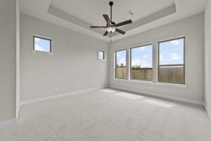 Empty bedroom with beige carpet, light gray walls, tray ceiling, ceiling fan with lights, and three large windows letting in natural light—perfect for those seeking painted tree homes for sale in McKinney, Texas.