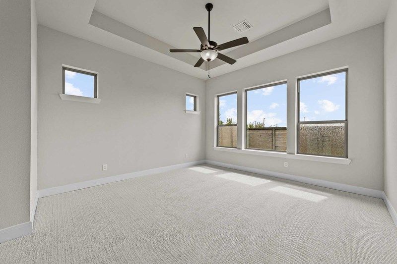 Empty bedroom with beige carpet, light gray walls, tray ceiling, ceiling fan with lights, and three large windows letting in natural light—perfect for those seeking painted tree homes for sale in McKinney, Texas.