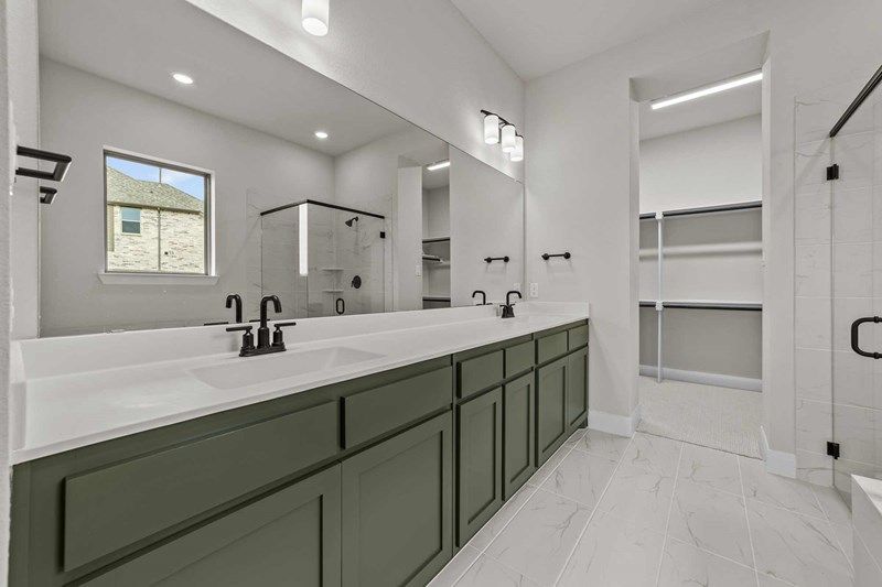 Modern bathroom in Painted Tree McKinney with green double-sink vanity, large mirror, black fixtures, and a walk-in closet. White tile floors and neutral walls complete this stylish space found in homes for sale in McKinney Texas.