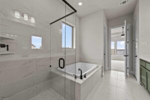 Modern bathroom with glass-enclosed shower, built-in bathtub, white tile walls and floors, large window, and black fixtures—showcasing the stylish design often found in homes for sale in McKinney, Texas. Doorway leads to a bright adjacent room.