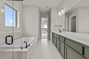 Modern bathroom with a glass shower, built-in bathtub, long green vanity with double sinks, and large mirror; door opens to a bright adjoining room—discover similar luxury in homes for sale in McKinney Texas.