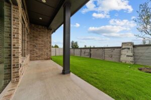 A covered patio with a concrete floor overlooks a grassy backyard enclosed by a tall wooden fence and a stone structure—an inviting setting found in many Painted Tree homes for sale in McKinney, Texas, under a partly cloudy sky.