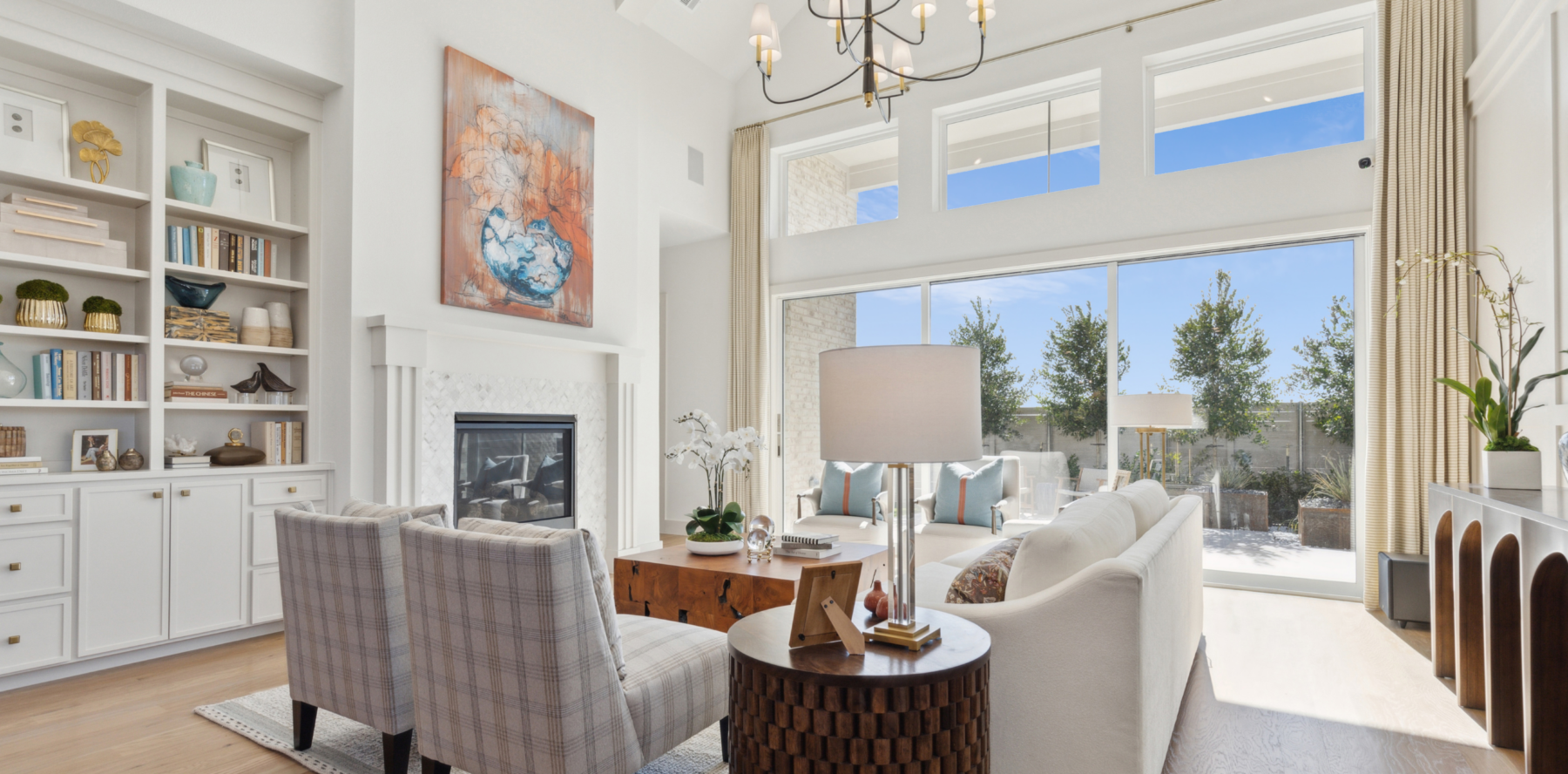 Bright living room in a Southgate Homes property, featuring white walls, a cozy fireplace, bookshelves, two plaid chairs, and a white sofa. A large window offers views of the TX patio with trees. Perfect for those seeking new homes with charm and comfort.