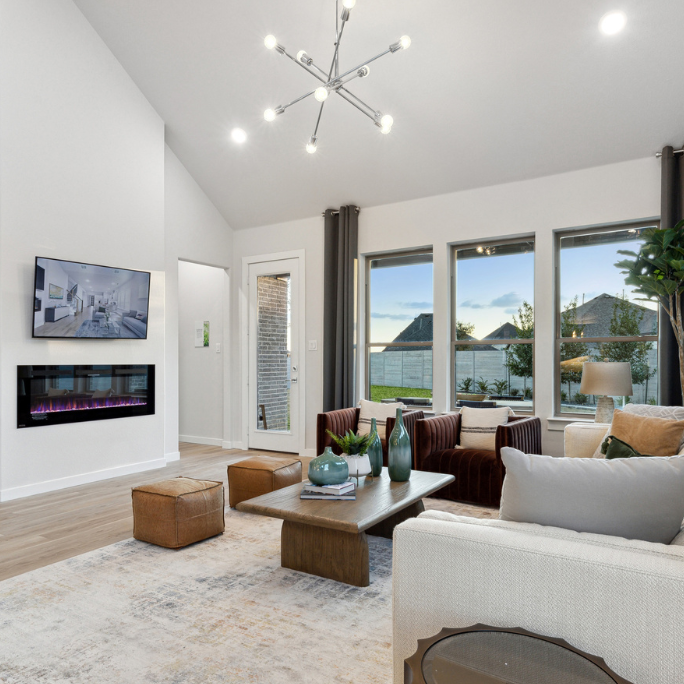 Discover a modern living room in one of McKinney's new homes at Painted Tree, featuring a beige sofa, brown chairs, and a wooden table. The wall-mounted TV and electric fireplace add warmth, while large windows and geometric ceiling lights enhance the airy space adorned with plants and ceramic vases.