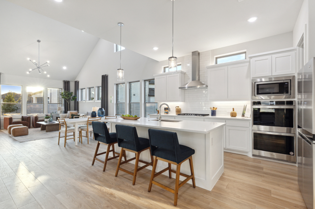 Modern open-concept kitchen and living room in a Trophy Signature Home, featuring high ceilings, white cabinets, an island with bar stools, dining table, and natural light from large windows. This exquisite space embodies the refined elegance found in McKinney's finest TX residences.