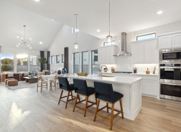 Modern kitchen and living area in one of McKinney's stunning new homes, featuring white cabinetry, island with bar stools, stainless steel appliances, and wooden flooring. Large windows provide natural light reminiscent of the serene beauty at Painted Tree.