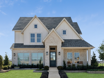 This two-story Southgate Home in McKinney features a brick facade, gray roof, and large front windows. A small walkway leads to the entrance, surrounded by manicured shrubs and a lush green lawn under a clear blue sky.