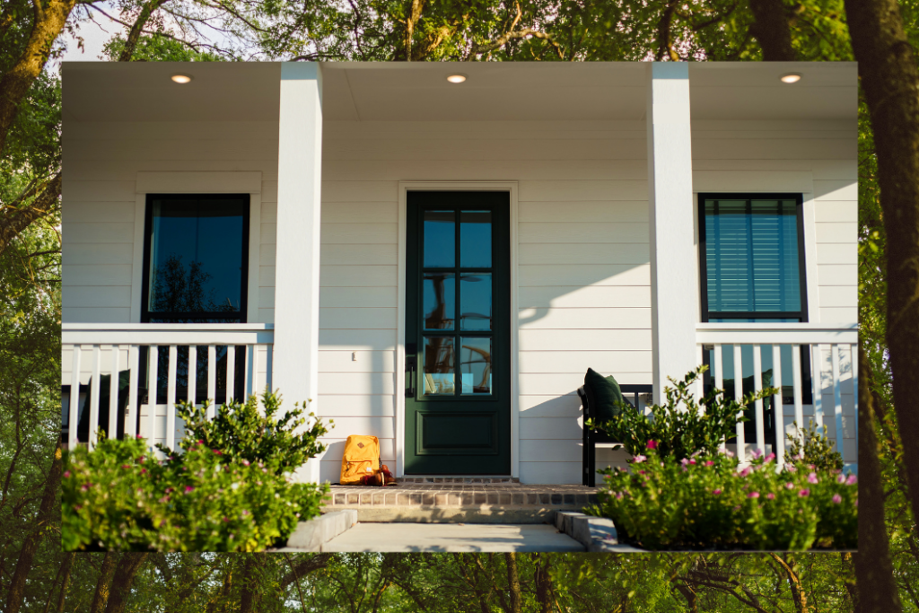 A top-selling community gem, this white house with a dark green door and two windows is embraced by woods and water. Steps lead to the porch, where a yellow backpack rests beside a bench among the trees.