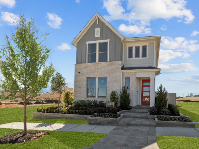 This stylish modern two-story house, built by David Weekley, features light brick and gray siding, a vibrant red front door, and a beautifully landscaped yard—all under a clear blue sky.