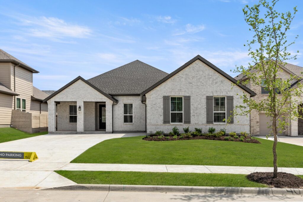 Single-story modern brick house with a manicured lawn, young tree, wide driveway, and two-car garage under a clear blue sky in Painted Tree McKinney—perfect for those seeking homes for sale in McKinney Texas.