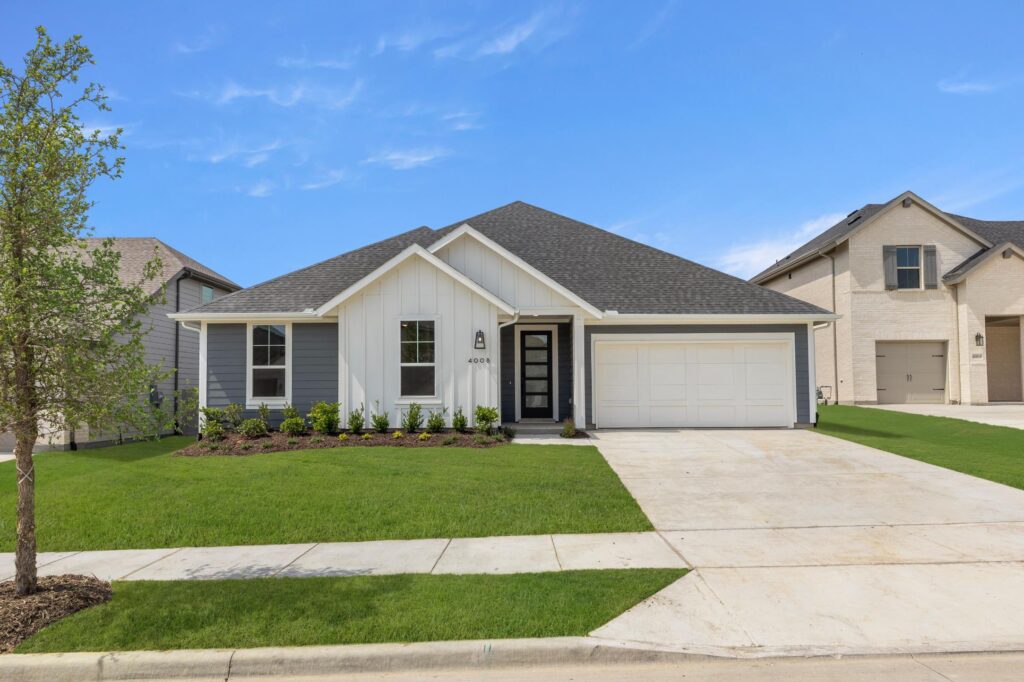 Charming single-story white house in Painted Tree McKinney with a dark gray roof, attached two-car garage, front porch, and well-maintained lawn on a suburban street under a blue sky. Explore Painted Tree homes for sale today!.