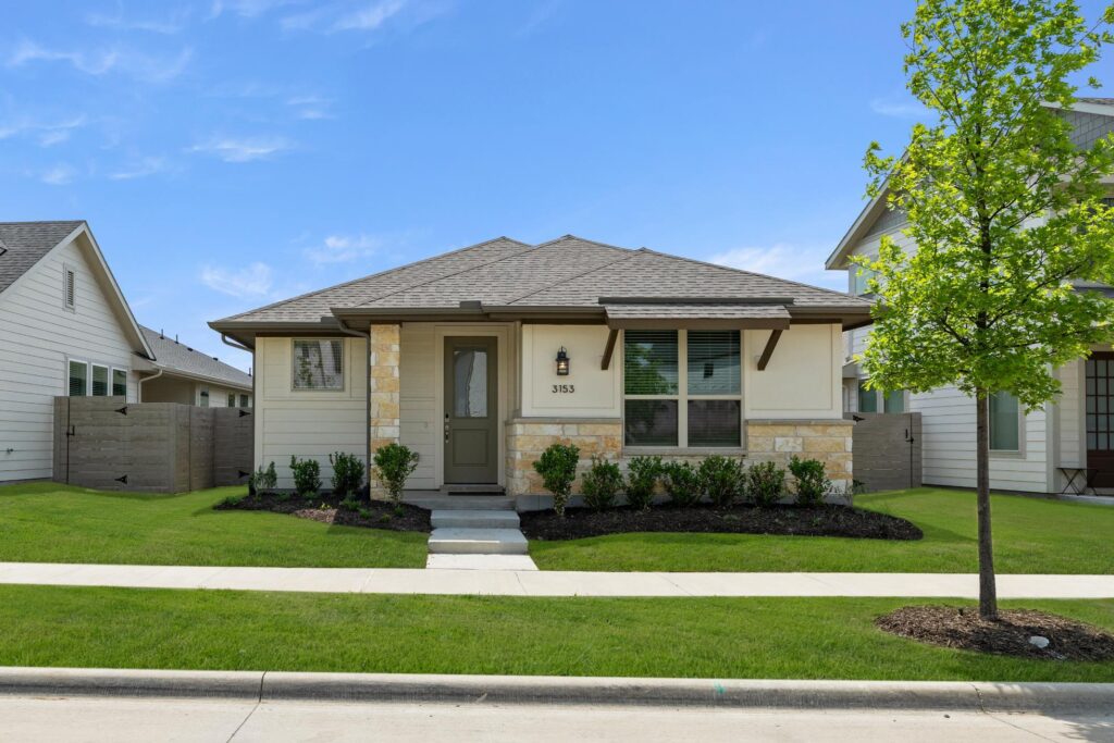 Single-story house with beige siding, stone accents, and a small front porch. The yard has green grass, shrubs, and a young tree near the sidewalk under a blue sky—perfect for those seeking Painted Tree homes for sale in McKinney, Texas.