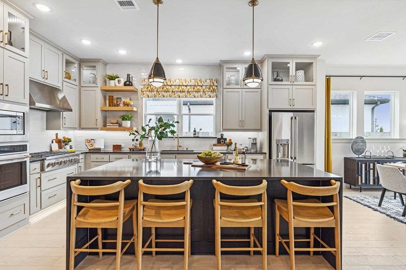 Modern kitchen with a large island, four wooden barstools, stainless steel appliances, pendant lights, and open shelves with decor—perfect for those seeking Painted Tree McKinney homes for sale. Dining area visible in the background.