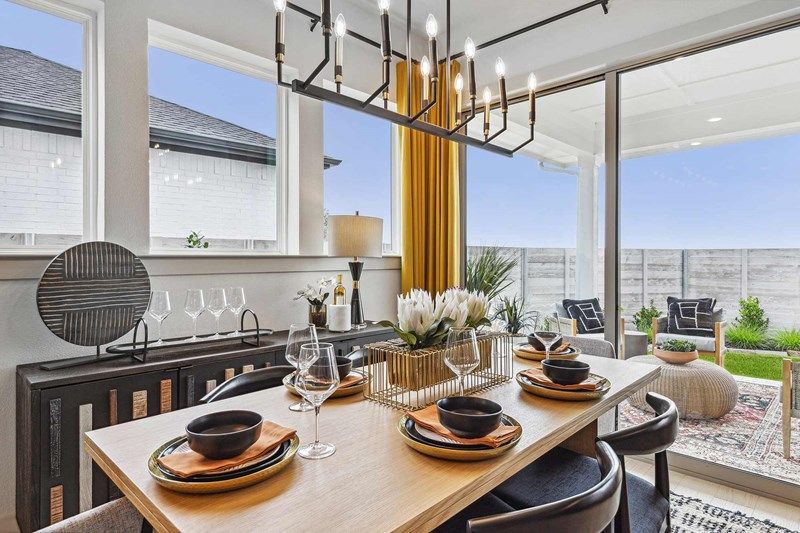 Modern dining room with a set table for four, large windows, gold curtains, chandelier, and a view of an outdoor patio—perfect for those seeking homes for sale in McKinney Texas or exploring Painted Tree TX.