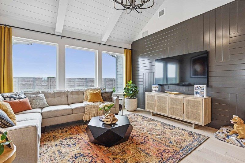 Modern living room in Painted Tree McKinney with a sectional sofa, patterned rug, black geometric coffee table, TV on a dark accent wall, and large windows with yellow curtains letting in natural light. Perfect for homes for sale in McKinney Texas.