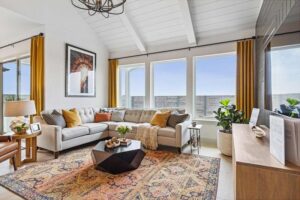 Modern living room in Painted Tree McKinney with a light sectional sofa, colorful rug, yellow curtains, large windows, lush indoor plants, and a wooden media console under vaulted white ceilings—perfect for homes for sale in McKinney Texas.