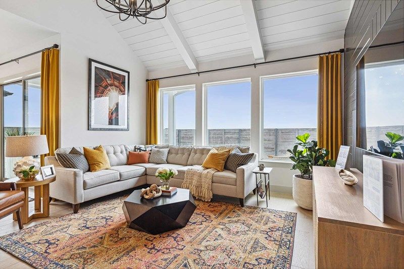 Modern living room in Painted Tree McKinney with a light sectional sofa, colorful rug, yellow curtains, large windows, lush indoor plants, and a wooden media console under vaulted white ceilings—perfect for homes for sale in McKinney Texas.