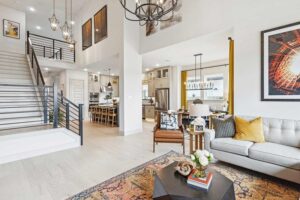 Modern open-concept living room with a gray sofa, armchair, and coffee table, adjacent to a kitchen and dining area, featuring a staircase and large wall art—ideal for those seeking Painted Tree homes for sale in McKinney.