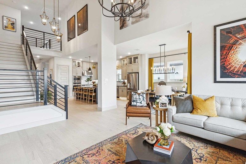 Modern open-concept living room with a gray sofa, armchair, and coffee table, adjacent to a kitchen and dining area, featuring a staircase and large wall art—ideal for those seeking Painted Tree homes for sale in McKinney.