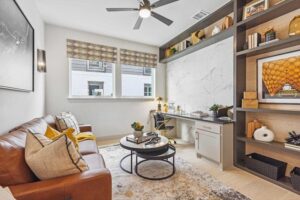 Modern home office with a brown leather sofa, round coffee table, built-in desk, shelves with decor, and two windows with patterned shades—perfect for those seeking Painted Tree McKinney style or homes for sale in McKinney, Texas.