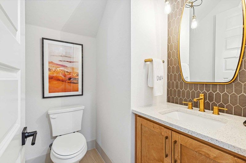 Modern bathroom in a Painted Tree McKinney home, featuring a toilet, wooden vanity, gold fixtures, hexagonal tile backsplash, and framed landscape artwork on the wall.
