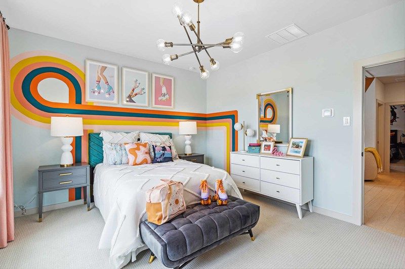 A modern bedroom in Painted Tree McKinney features a retro mural, colorful bedding, a gray bench with stuffed toys, and a dresser with framed photos and a mirror. Discover stylish spaces like this in homes for sale in McKinney Texas.