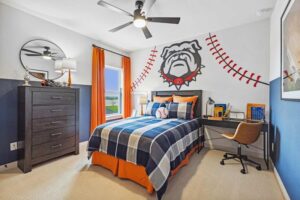 A sports-themed bedroom in the heart of Painted Tree McKinney features a plaid bed, orange accents, desk, and a mural with a baseball and bulldog mascot—perfect inspiration for those exploring homes for sale in McKinney Texas.