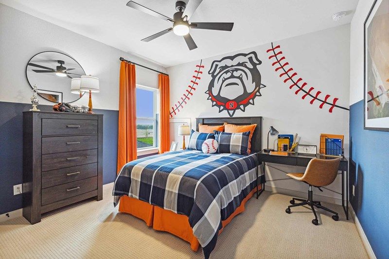 A sports-themed bedroom in the heart of Painted Tree McKinney features a plaid bed, orange accents, desk, and a mural with a baseball and bulldog mascot—perfect inspiration for those exploring homes for sale in McKinney Texas.